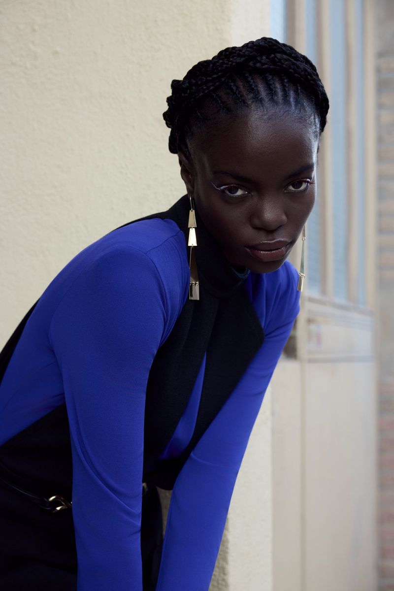 A stylish portrait of a Black woman with braided hair wearing a blue top and black vest with gold earrings, looking directly at the camera.
