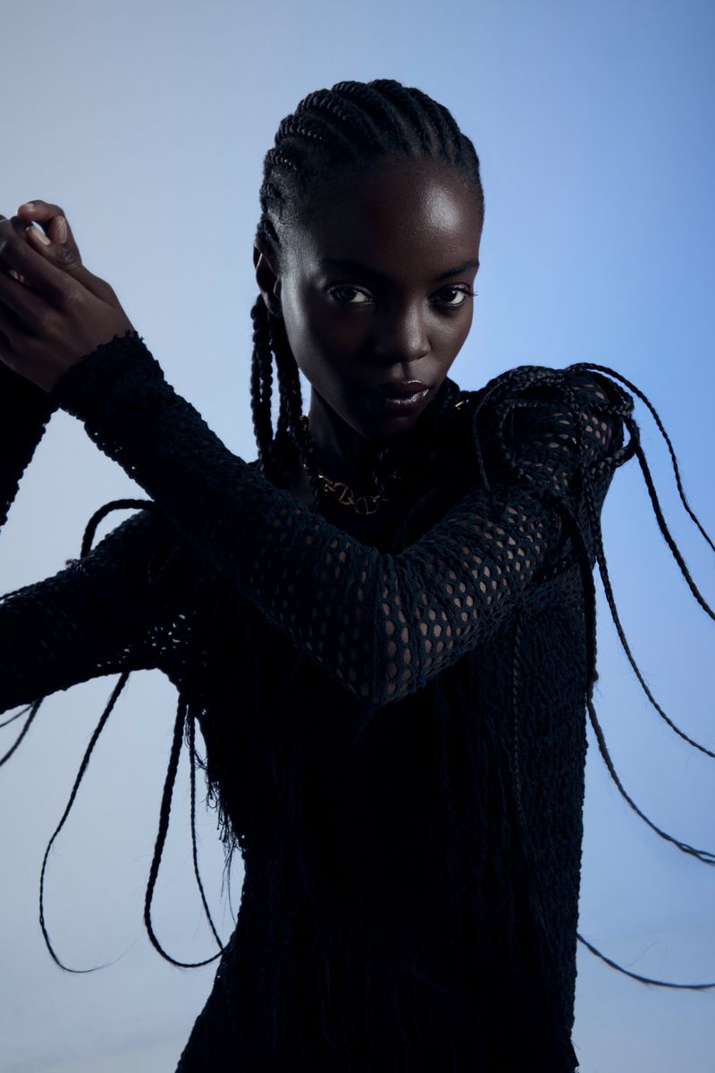 Portrait of a black female fashion model with braided hair, wearing a black knit top against a light blue studio background.