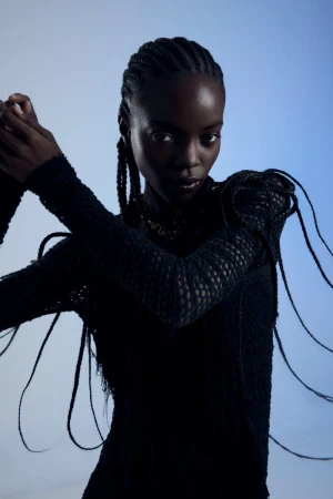 Portrait of a black female fashion model with braided hair, wearing a black knit top against a light blue studio background.
