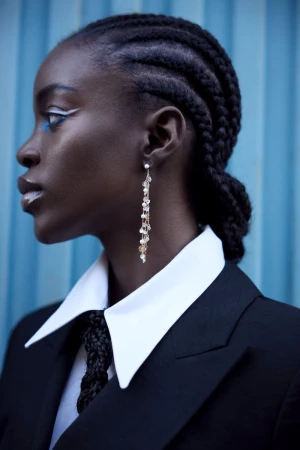 Profile portrait of a Black woman with cornrows, wearing a black blazer with white collared shirt, and ornate earrings, against blue backdrop.