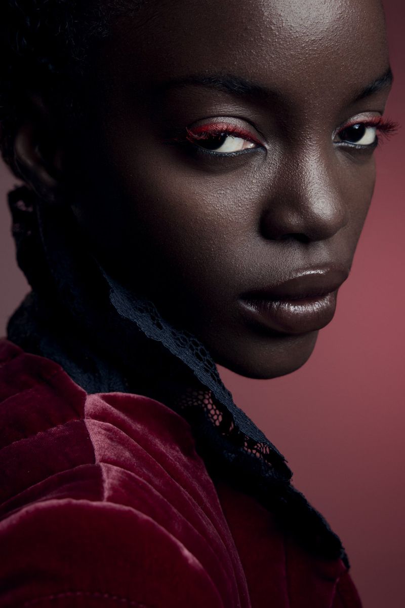 Close-up portrait of a Black woman with dark eyes and red eye makeup, wearing a red velvet jacket with lace collar on red backdrop.