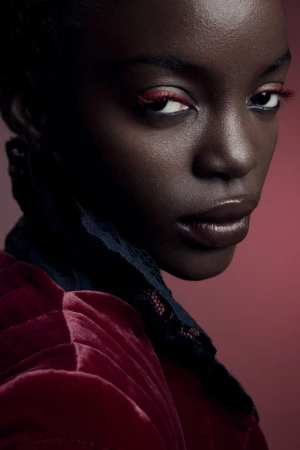 Close-up portrait of a Black woman with dark eyes and red eye makeup, wearing a red velvet jacket with lace collar on red backdrop.