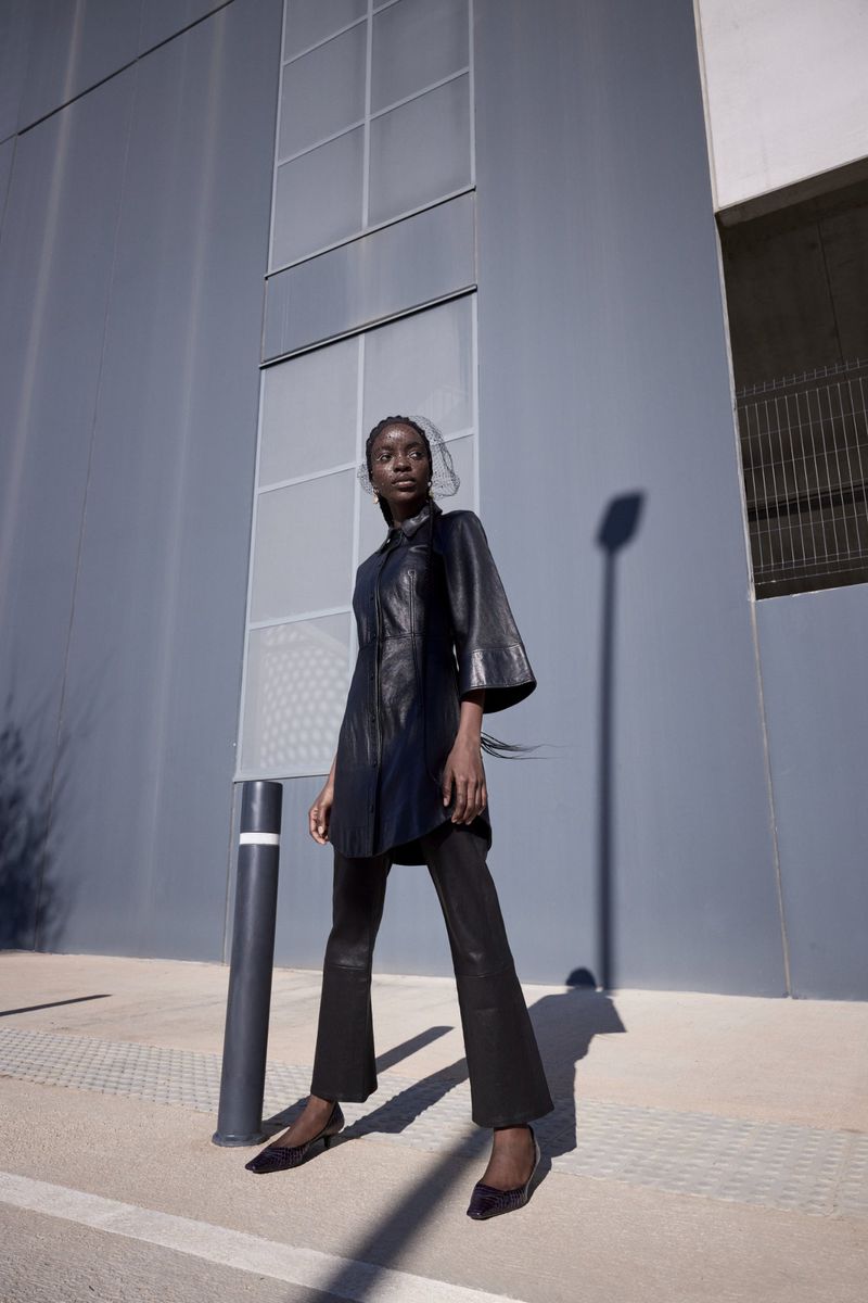 Fashion photo of a Black woman wearing a black leather outfit and flats, posing outdoors against a grey modern architectural wall.
