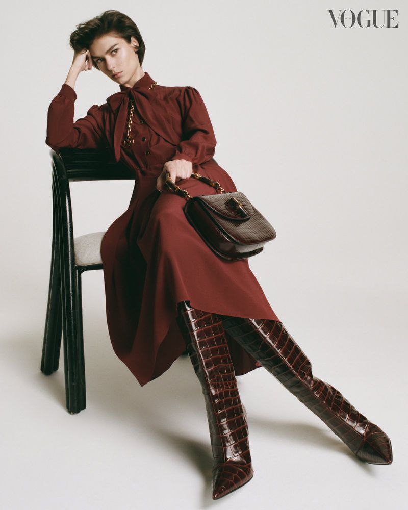 Fashion editorial shot of a woman in a dress and crocodile-print boots sitting on a chair, holding a purse in a neutral-toned studio.