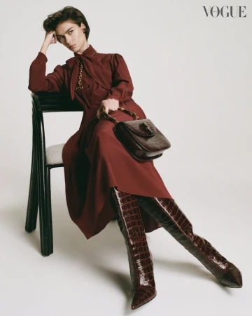 Fashion editorial shot of a woman in a dress and crocodile-print boots sitting on a chair, holding a purse in a neutral-toned studio.