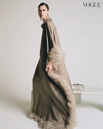 Fashion editorial shot of a female model wearing a patterned coat with feather trim, posing in a photo studio with a handbag.