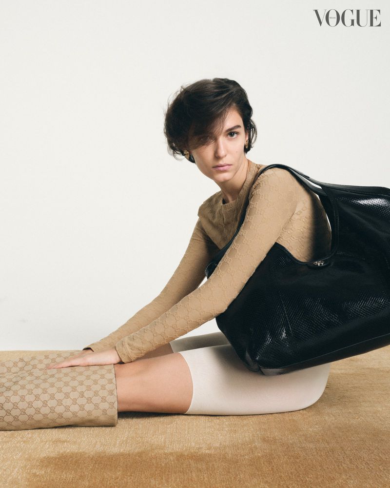 A woman poses with a black shoulder bag in a studio setting, wearing a tan long-sleeve shirt and patterned leggings. Vogue logo visible.