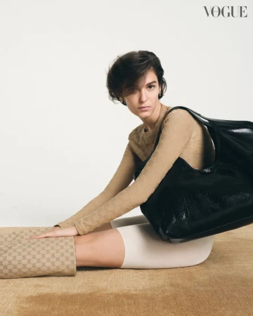 A woman poses with a black shoulder bag in a studio setting, wearing a tan long-sleeve shirt and patterned leggings. Vogue logo visible.