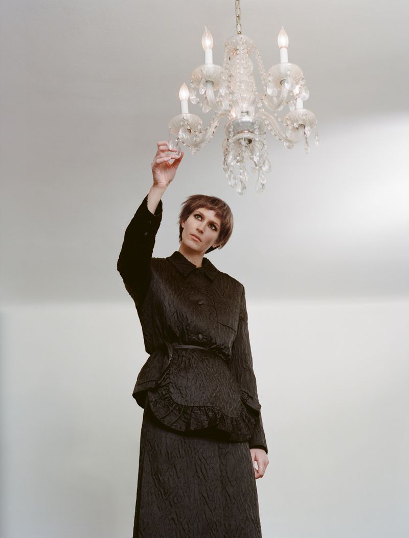 A woman in a black suit stands indoors reaching toward a lit crystal chandelier against a plain, light background, in a fashion context.