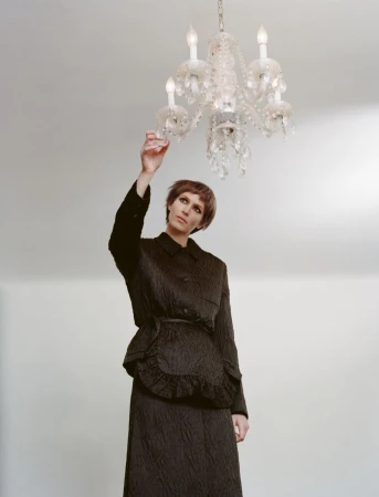 A woman in a black suit stands indoors reaching toward a lit crystal chandelier against a plain, light background, in a fashion context.