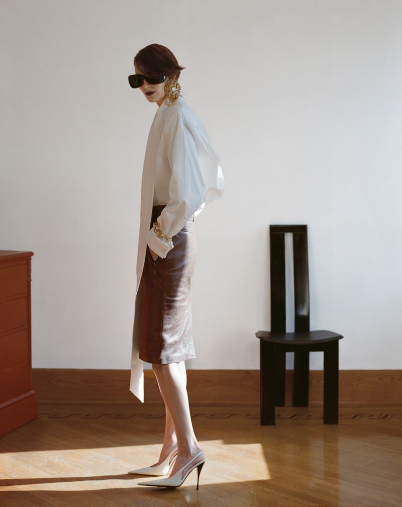 Elegant woman in a white blouse and brown pencil skirt posing indoors with a black chair against a neutral backdrop and wood floor.
