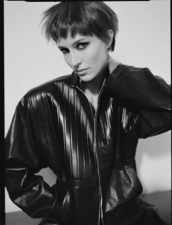 Monochrome portrait of a woman with short hair in a black leather jumpsuit posing against a white wall, showcasing fashion and style.