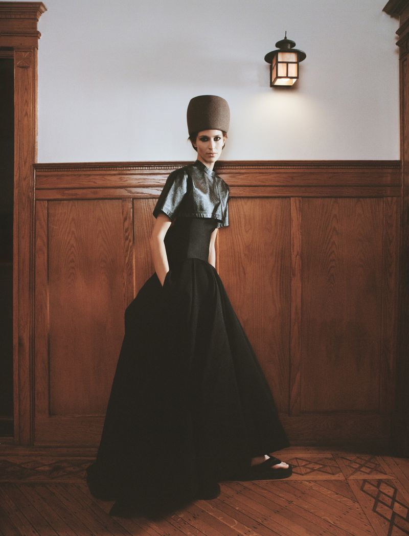 A woman models a dramatic black gown and matching cylindrical hat indoors, posing against wood paneling and a white wall with a lantern.