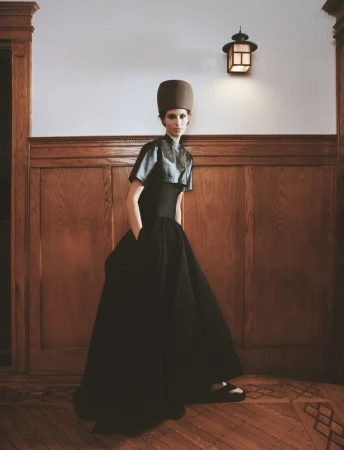 A woman models a dramatic black gown and matching cylindrical hat indoors, posing against wood paneling and a white wall with a lantern.