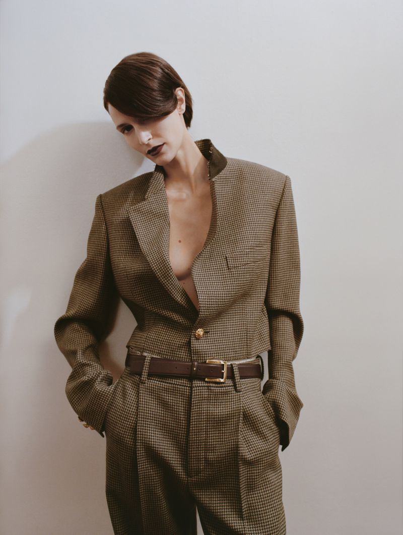 A woman in a houndstooth suit with a brown belt poses against a light-colored wall, shot in portrait orientation with a studio lighting.