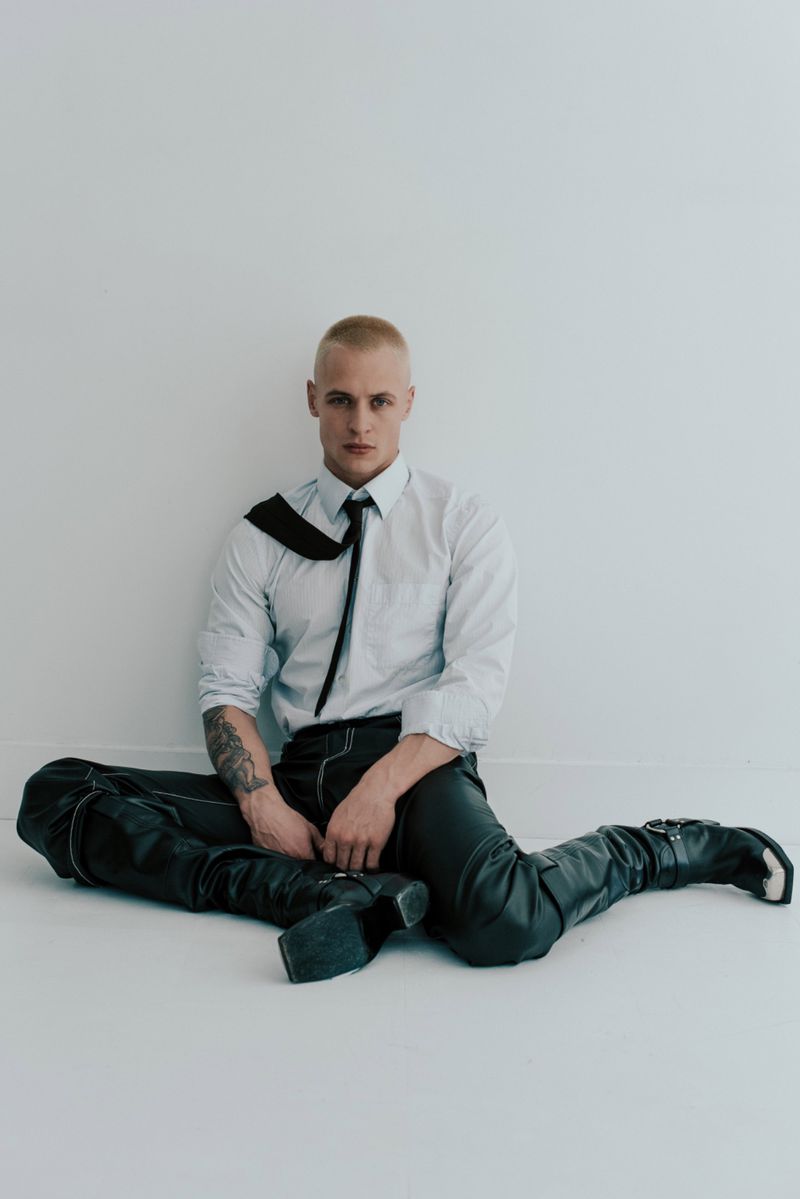 Studio shot of a young adult man with a buzzcut, wearing an unbuttoned shirt, tie, leather pants and boots, sitting on a white floor.