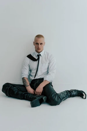 Studio shot of a young adult man with a buzzcut, wearing an unbuttoned shirt, tie, leather pants and boots, sitting on a white floor.