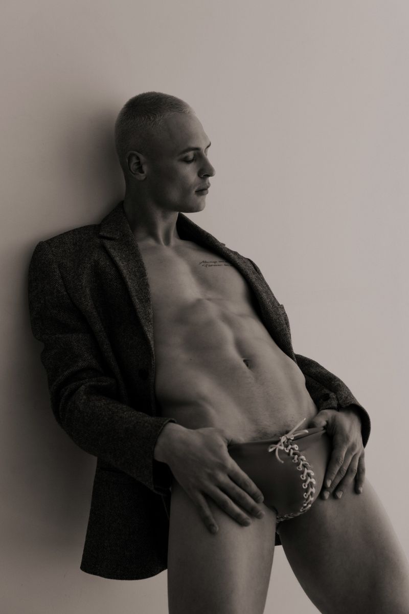 Monochrome portrait of a blond man wearing a tweed blazer with bare torso and posing in briefs indoors against a plain wall.