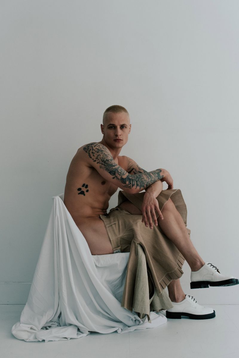 Tattooed man with buzzcut sits in photo studio wearing khaki skirt and white shoes, draped with fabric, against a blank background.