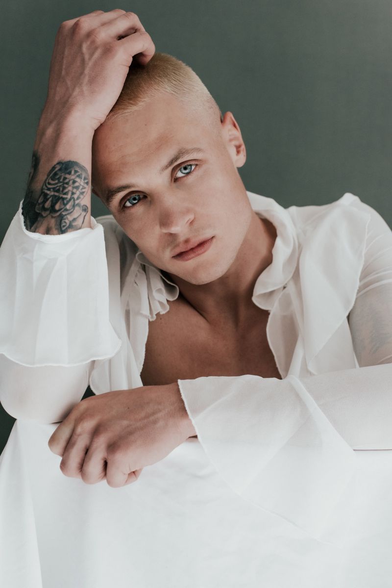 Portrait of a blond male model in a white ruffled shirt, with a tattoo on his arm, resting his head on his hand in a photo studio.