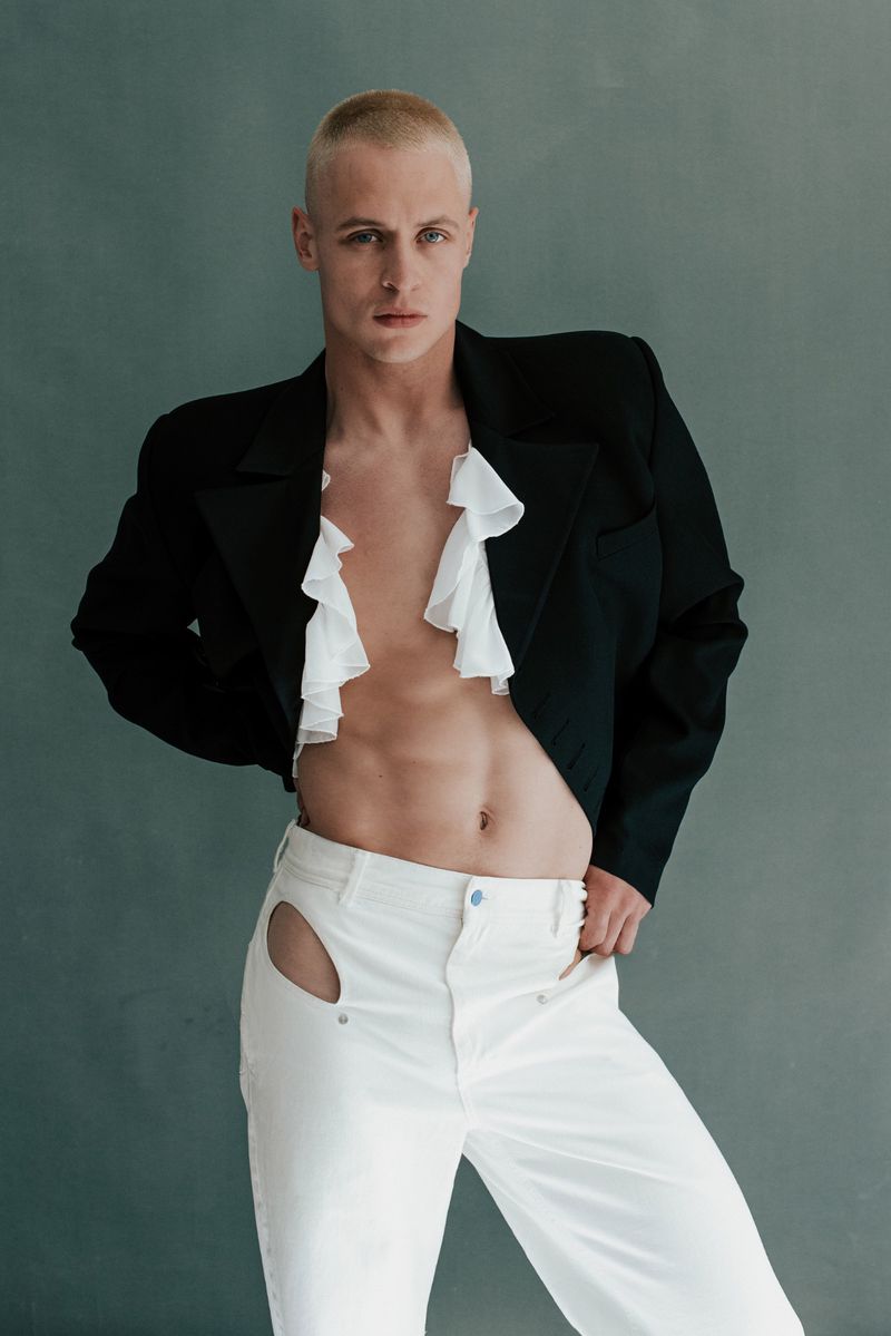 Man with a shaved head poses in a black jacket, ruffled white top, and white pants with cutouts, looking directly at the camera against gray backdrop.