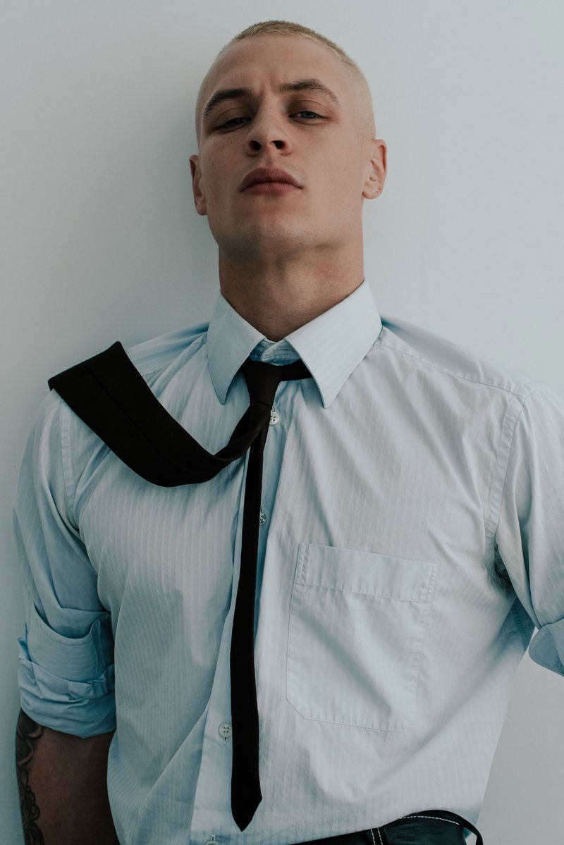 Studio shot of an adult male wearing a blue dress shirt and dark necktie against a plain light grey background with natural light.