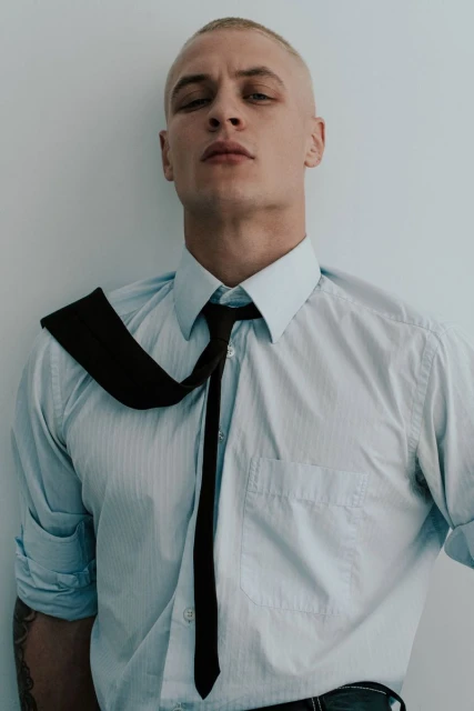 Studio shot of an adult male wearing a blue dress shirt and dark necktie against a plain light grey background with natural light.