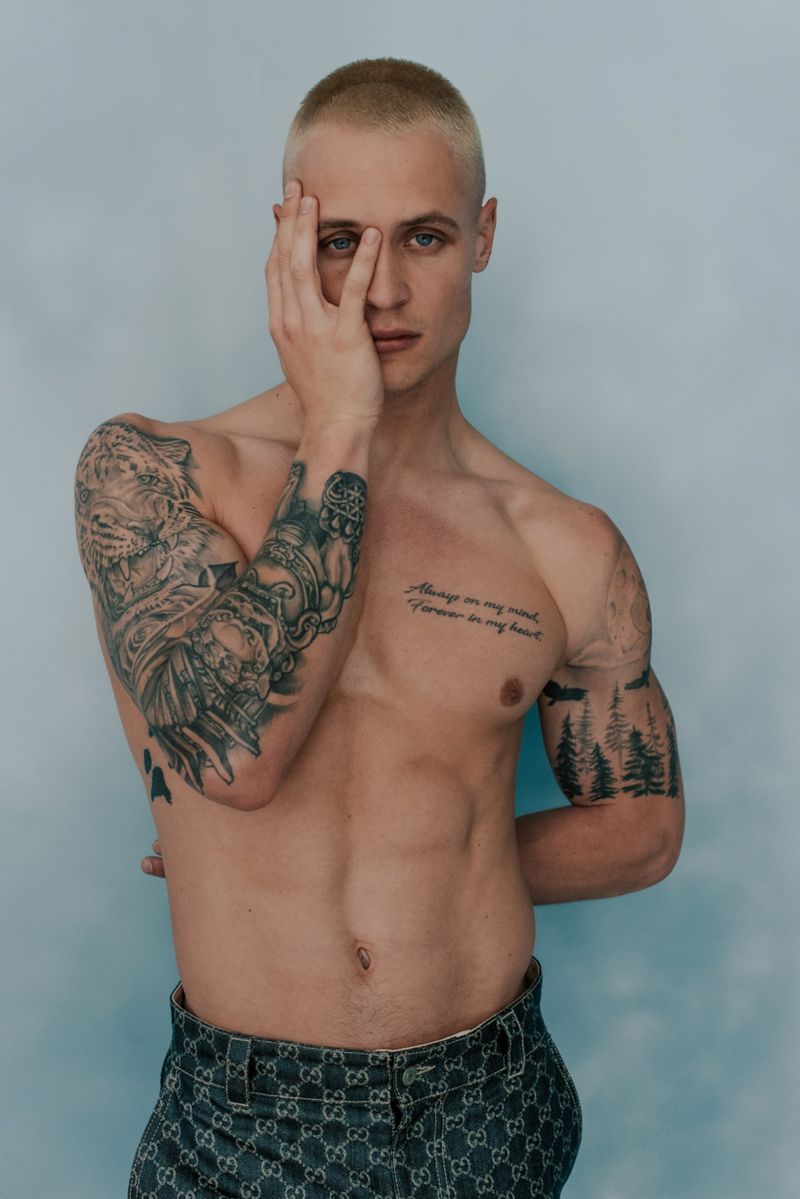 Shirtless man with tattoos poses with hand on face in studio with light blue background. Fashion and beauty portrait photography.