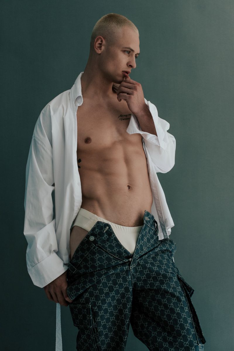 Studio shot of a blond, muscular, shirtless man in loose unbuttoned shirt and patterned pants striking a contemplative pose.