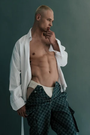 Studio shot of a blond, muscular, shirtless man in loose unbuttoned shirt and patterned pants striking a contemplative pose.