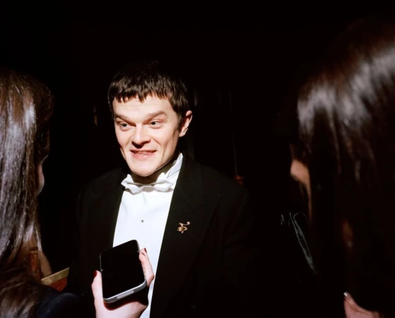 A man in a tuxedo is surrounded by two women, holding a cell phone. He is at an event with a dark background, indoors, with event lighting.