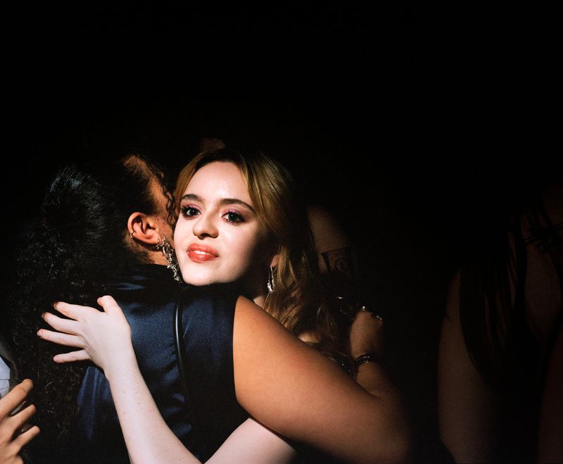 Two women embrace in dimly lit setting, one with auburn hair and red lipstick, the other with dark curly hair, both wearing cocktail dresses.
