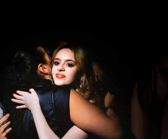 Two women embrace in dimly lit setting, one with auburn hair and red lipstick, the other with dark curly hair, both wearing cocktail dresses.