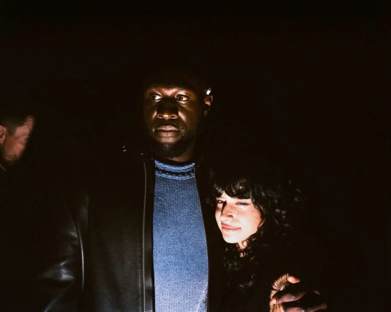 A man and a woman pose indoors with a second man visible in the background. The image is lit with a warm light source and is in portrait orientation.