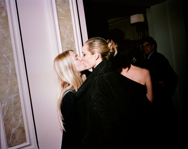 Two adult females kissing indoors near a doorway, other partygoers in the dark background. Soft lighting and candid camera style.
