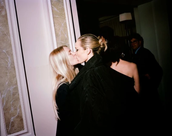 Two adult females kissing indoors near a doorway, other partygoers in the dark background. Soft lighting and candid camera style.