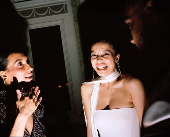 Three individuals laughing indoors at a party, illuminated by shadows and bright light on their faces and stylish attire.