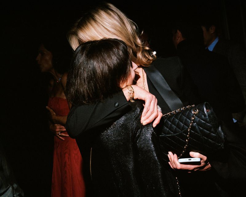 Two women embrace in a dimly lit setting, one with blonde hair and a black blazer, the other with short dark hair and a textured top.
