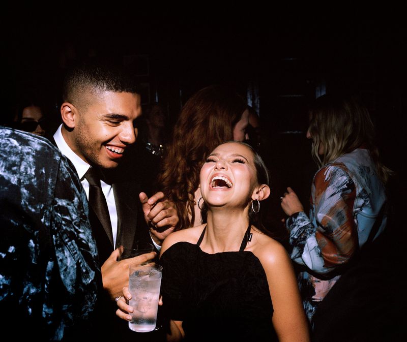 Group of young adults laugh in dim, indoor light. Woman in foreground holds drink while smiling. Focus on people, indoor event.