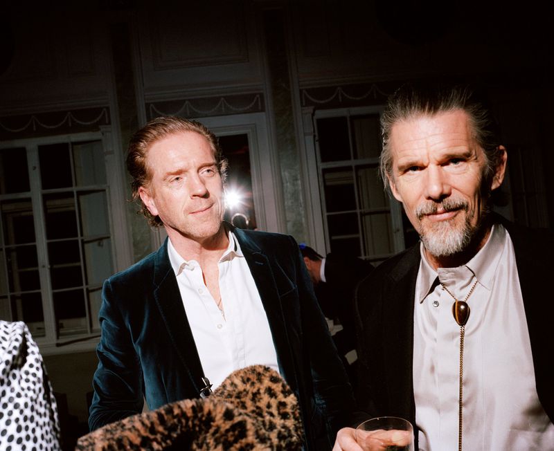 Two adult males pose indoors in formal wear. One man wears a velvet jacket, while the other sports a bolo tie and holds a drink.