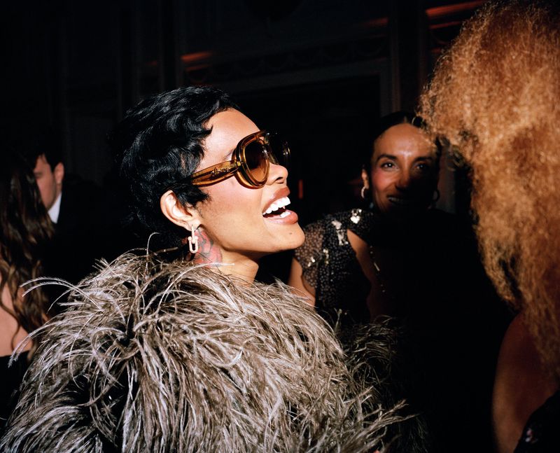 Elegant woman in sunglasses and feather boa smiles at a party, surrounded by dark background and other attendees.