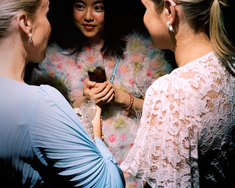 Three stylish women gather at an event, showcasing elegant dresses, drinks, and radiant smiles in a softly lit, intimate setting.
