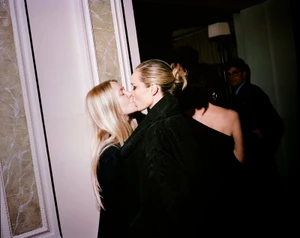 Two women kissing in a dimly lit interior space with ornate architectural columns and background figures.