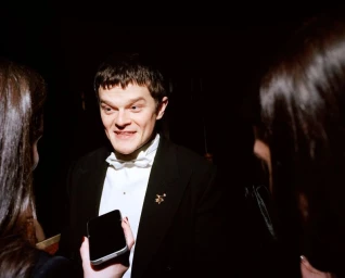 Man in a tuxedo with a white bow tie surrounded by people holding a smartphone up in a dark setting