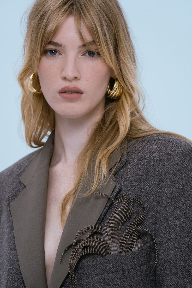 Portrait of a woman with long blonde hair and gold hoop earrings wearing a grey jacket with a decorative feather in the pocket.