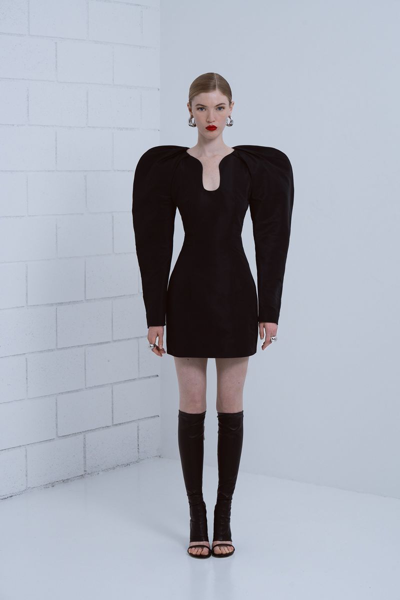 Elegant red-headed woman in black mini dress with puffed shoulders and knee-high boots posing against a white brick wall.