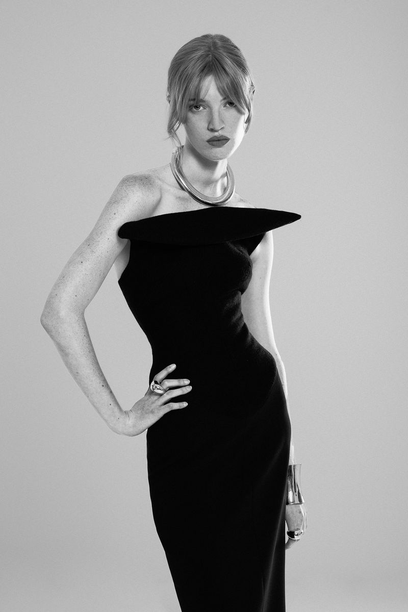 Black and white portrait of a red-haired woman in a black strapless dress, wearing silver jewelry and posing in a photo studio.
