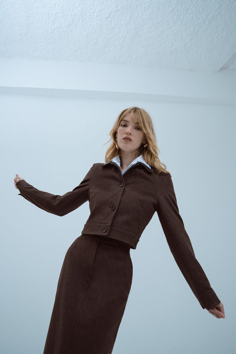 A woman models a brown skirt suit with a collared white shirt, posed in an indoor space with angled lighting and a low-angle perspective.
