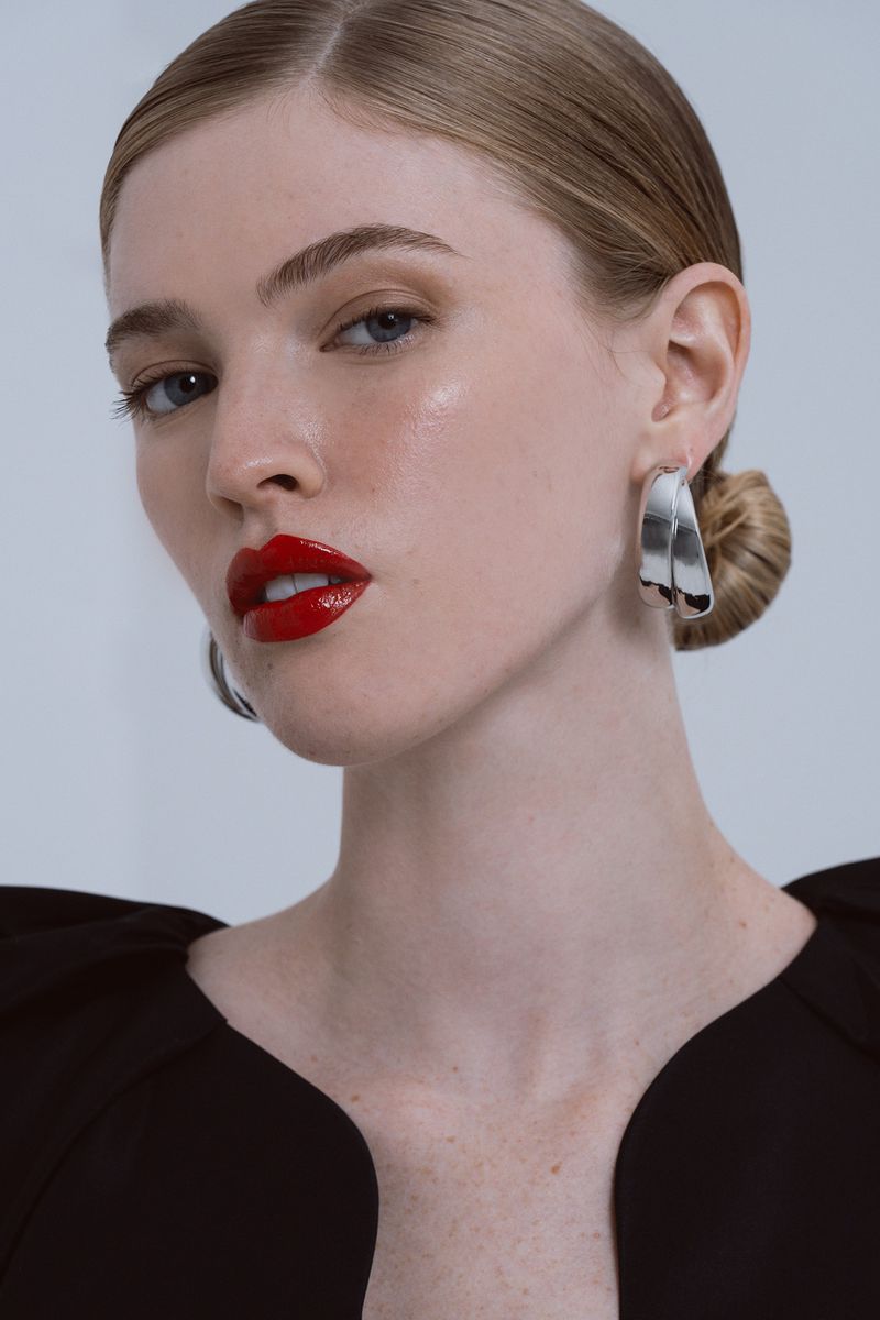 Close-up portrait of a woman with red lipstick and statement earrings in a studio. She has fair skin, light eyes, and styled blonde hair.