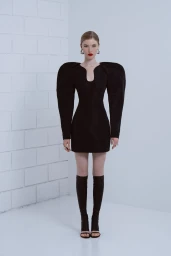 Model in a black structured minidress with exaggerated shoulders and tall boots standing against a white brick wall